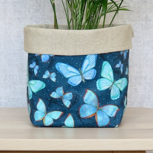 May include: A blue fabric planter with a light brown linen top. The planter features a repeating pattern of blue butterflies on a dark blue background with white polka dots.