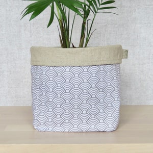 May include: A white planter with a gray geometric pattern of overlapping semicircles. The planter has a natural linen top and is filled with a green plant.