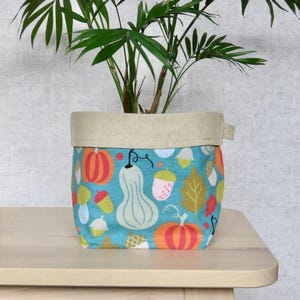 May include: A teal fabric planter with a beige linen top. The planter features a colourful pattern of pumpkins, gourds, acorns, and leaves.