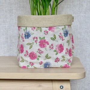 May include: A fabric planter with a floral pattern of pink and blue flowers on a white background. The top edge is a neutral beige colour. The planter is on a light wood surface and contains green plants.