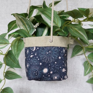 May include: A hanging planter made of natural linen fabric with a dark blue print of celestial bodies, including stars, moons, and suns. The planter is filled with a green plant with white stripes on the leaves.