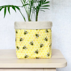 May include: A yellow fabric planter with a honeycomb pattern and a repeating image of bees. The planter has a beige linen top and is filled with a green plant.