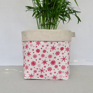 May include: A square fabric plant pot with a red snowflake pattern on a white background. The top edge is a natural linen colour. The pot contains a green plant.