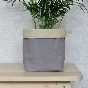 May include: A grey fabric planter with a beige linen top. The planter is sitting on a light wood surface and has a small tag with the word "Handmade" on it.