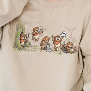 May include: Beige crewneck sweatshirt featuring a whimsical illustration of Ewoks in a forest setting. The artwork depicts several Ewoks interacting with a Stormtrooper helmet. The design is detailed and colorful, perfect for fans of the Star Wars franchise.