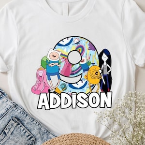 May include: White t-shirt featuring a colorful graphic of cartoon characters and the name "Addison" in black. The design includes characters from a popular animated series, with a number 9 in the background.