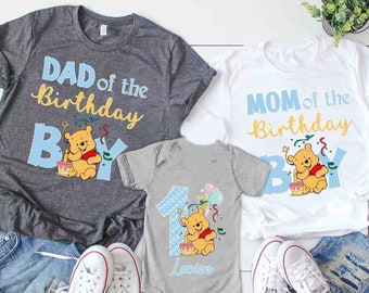 Puede incluir: Tres camisetas de cumpleaños a juego con el tema de Winnie the Pooh y un body. Las camisetas dicen "DAD of the Birthday Boy", "MOM of the Birthday Boy", y el body dice "1" con el nombre "Lucian". Todos presentan a Pooh con un pastel.