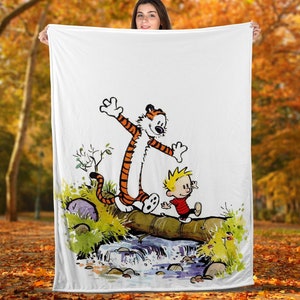 May include: White blanket featuring a cartoon illustration of Calvin and Hobbes. Hobbes, an orange tiger, stands on a log with Calvin, a young boy in a red shirt. The scene includes a stream, greenery, and a fall foliage background.