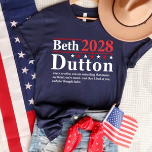 May include: Navy blue t-shirt with the text "Beth 2028 Dutton" in white and red. The shirt features a quote below the text. The image includes a hat, denim shorts, a red bandana, and an American flag.