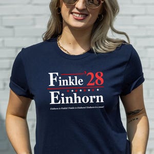 May include: Navy blue t-shirt with the text "Finkle '28 Einhorn" in white and red. The design includes five stars and the phrase "Einhorn is Finkle! Finkle is Einhorn! Einhorn is a man!"