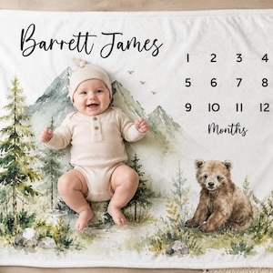 May include: A white milestone blanket featuring a watercolor mountain landscape, pine trees, and a brown bear cub. The text reads Barrett James and includes numbers 1 through 12 followed by the word Months for tracking a baby's age in months.