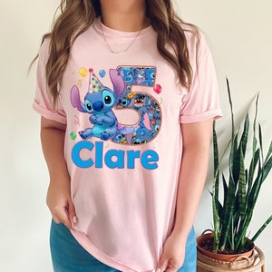May include: A light pink t-shirt featuring a cartoon character, Stitch, wearing a party hat. The number "5" is filled with Stitch images, and the name "Clare" is written in blue. Colorful balloons and confetti are also present.