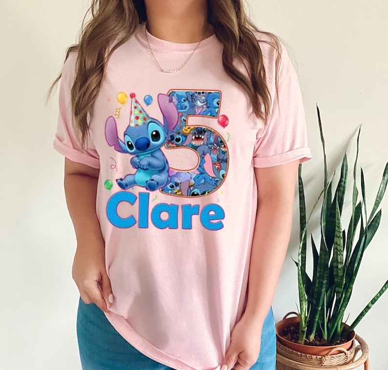 May include: A light pink t-shirt featuring a cartoon character, Stitch, wearing a party hat. The number "5" is filled with Stitch images, and the name "Clare" is written in blue. Colorful balloons and confetti are also present.