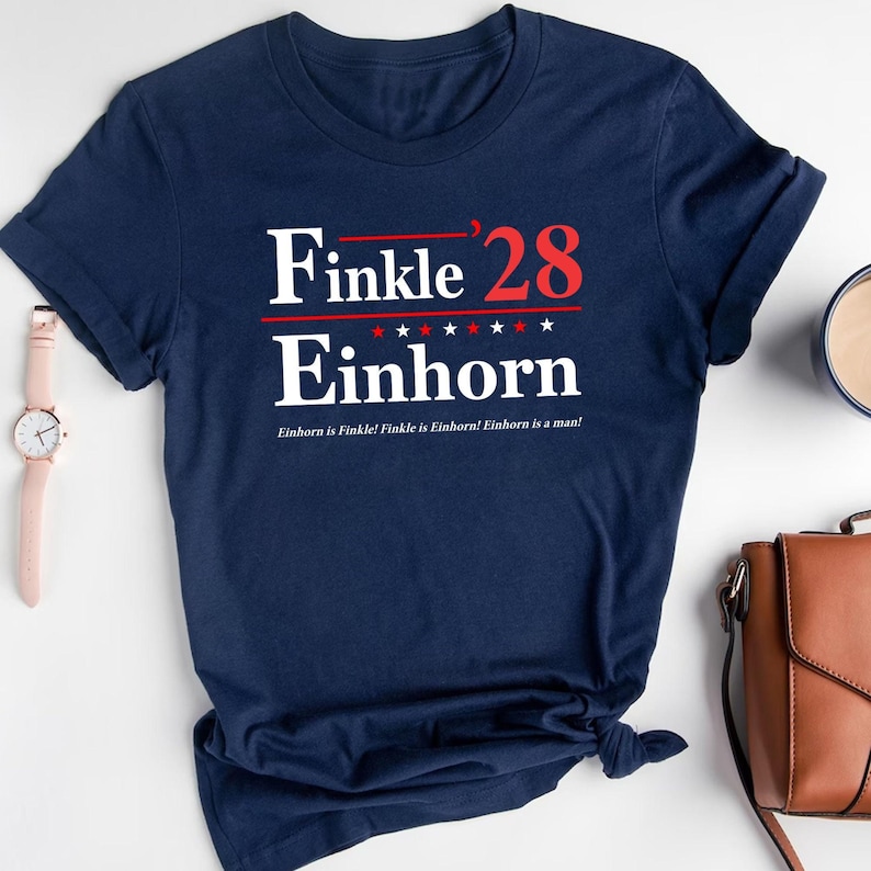 May include: Navy blue t-shirt with the text "Finkle '28 Einhorn" in white and red. The shirt also has the phrase "Einhorn is Finkle! Finkle is Einhorn! Einhorn is a man!" printed on it. The shirt is knotted at the bottom.