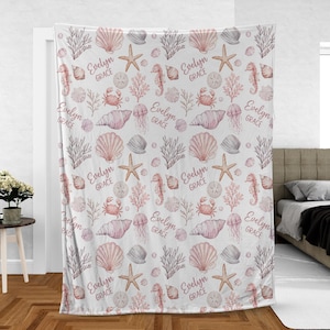 May include: A white fleece blanket featuring a repeating pattern of pink watercolour sea life including seashells, starfish, seahorses, crabs, jellyfish, and coral. The text Evelyn Grace is printed in a cursive script throughout the design.