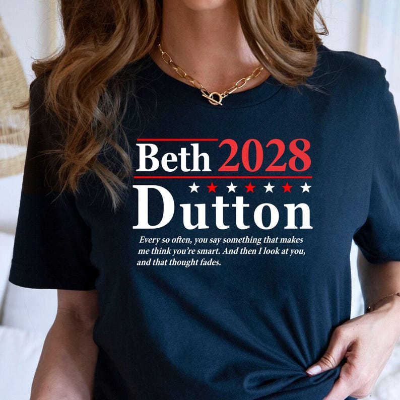 May include: Navy blue t-shirt with the name "Beth Dutton 2028" in white and red text. The shirt also features a quote in white text: "Every so often, you say something that makes me think you're smart. And then I look at you, and that thought fades."