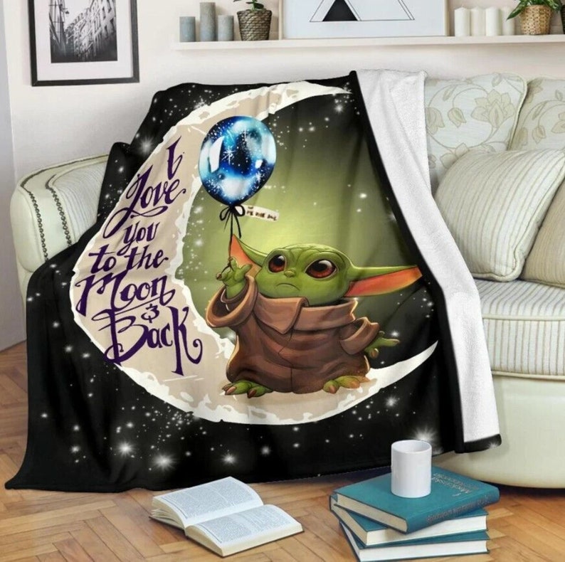 May include: A plush blanket featuring a green alien character holding a blue balloon against a crescent moon and starry background. The blanket has the text "I Love You to the Moon & Back" in a stylized font. The blanket is draped over a sofa.