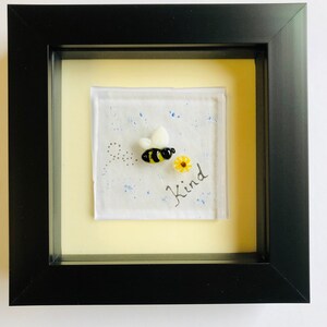 May include: A framed glass art piece with a yellow and black bee, a yellow flower, and the words "Be Kind" in black cursive writing.