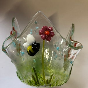 May include: A clear glass votive candle holder with a green, yellow, white, black, pink, and orange bee and flower design.