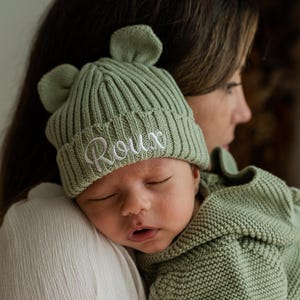 May include: A sage green ribbed knit beanie with two small rounded ears on top and the name Roux embroidered in white script on the folded cuff, paired with a matching sage green long-sleeved knit hooded romper with a textured stitch pattern.