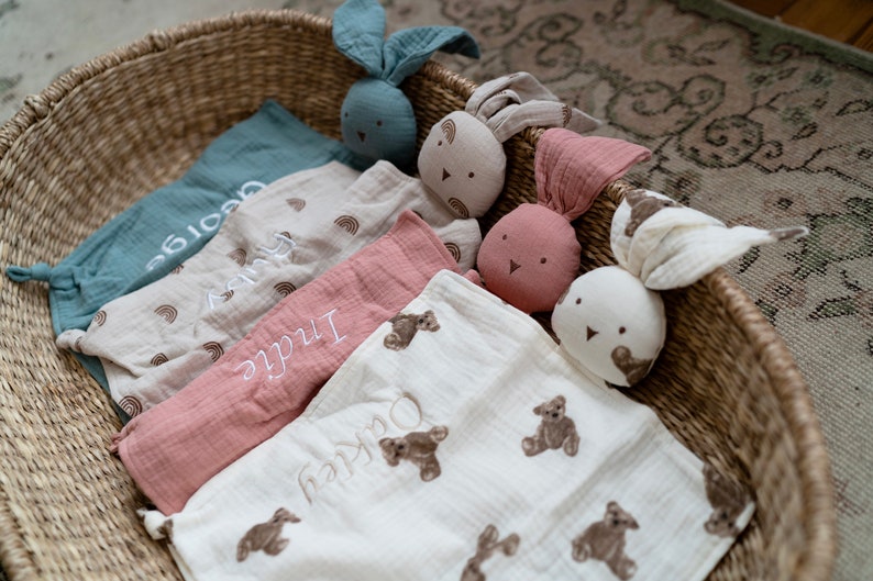 May include: Four personalized baby blankets in a wicker basket. The blankets are made of soft, muslin fabric and feature embroidered names and designs. The blankets are in shades of blue, beige, pink, and white. There are also three plush bunny toys in the basket.