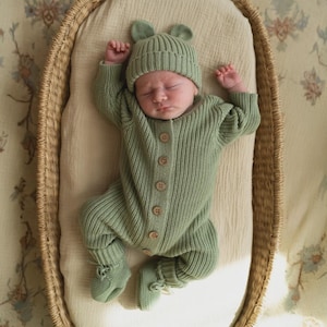 May include: A sage green knitted long-sleeve romper with a matching beanie featuring small bear ears. The romper has a vertical ribbed texture, a round neckline, and a front placket with six light brown wooden buttons. The outfit includes matching booties.