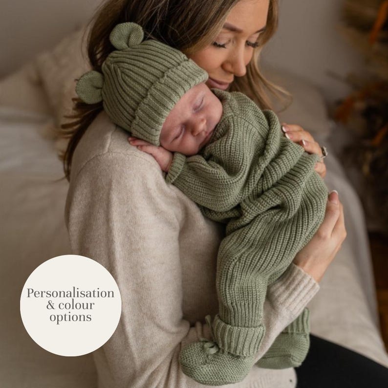 May include: A sage green ribbed knit baby outfit consisting of a long-sleeved romper with integrated footies and a matching beanie hat featuring two small rounded ears on top. The image includes a circular white graphic with the text Personalisation & colour options.