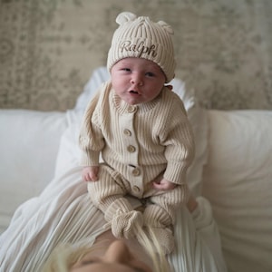 May include: A beige ribbed knit baby romper with long sleeves and matching booties, featuring a front button closure. The baby wears a matching beige ribbed knit beanie with small bear ears and the name Ralph embroidered in brown script.