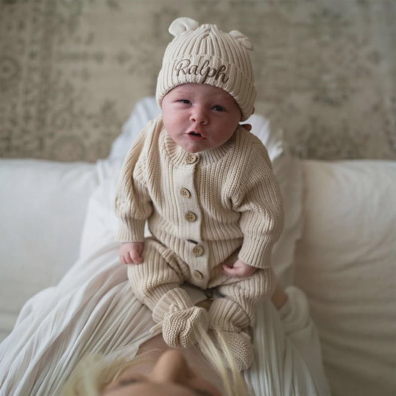 May include: A beige ribbed knit baby romper with long sleeves and matching booties, featuring a front button closure. The baby wears a matching beige ribbed knit beanie with small bear ears and the name Ralph embroidered in brown script.