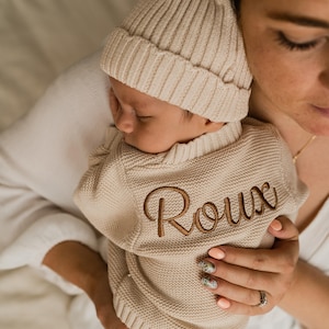 May include: A beige knitted baby sweater featuring the name Roux embroidered in brown script lettering on the chest, paired with a matching beige ribbed knit beanie hat. The garment has a textured knit pattern and a ribbed collar and hem.