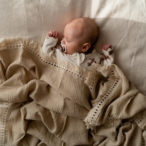 May include: A sleeping baby wearing a white onesie with a brown pattern, and a pacifier, is nestled under a beige blanket with a fringed edge. The blanket is draped over a light-colored surface.