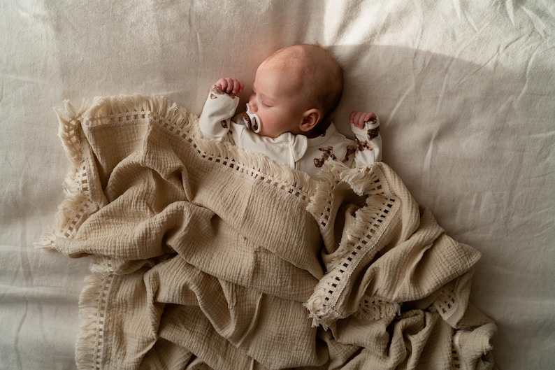 May include: A sleeping baby wearing a white onesie with a brown pattern, and a pacifier, is nestled under a beige blanket with a fringed edge. The blanket is draped over a light-colored surface.