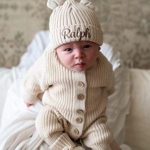 May include: A beige ribbed knit baby outfit consisting of a long-sleeved button-up romper, matching booties, and a beanie with small bear ears. The beanie features the name Ralph embroidered in brown script on the front cuff.