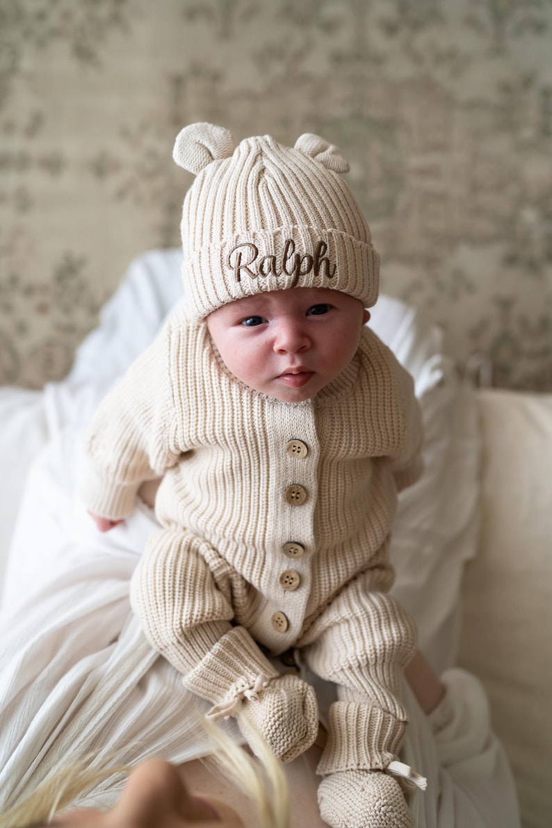 May include: A beige ribbed knit baby outfit consisting of a long-sleeved button-up romper, matching booties, and a beanie with small bear ears. The beanie features the name Ralph embroidered in brown script on the front cuff.