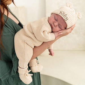 May include: A beige knitted baby outfit consisting of a long-sleeved romper, matching booties with ties, and a ribbed beanie with small ears. The beanie features the name Riley embroidered in brown script on the front of the knit fabric.