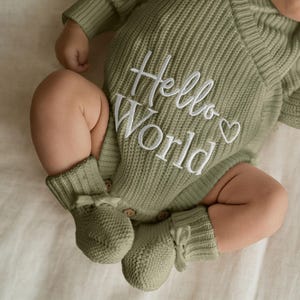 May include: A sage green knitted romper featuring a wrap-style neckline and white embroidered text that reads Hello World with a small heart icon. The garment includes wooden buttons at the crotch and matching sage green knitted booties with bows.