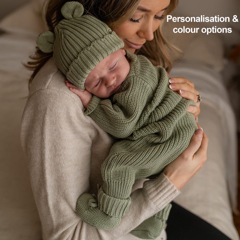 May include: A sage green knitted baby outfit consisting of a ribbed beanie with small bear ears, a long-sleeved sweater, matching pants, and booties. The text "Personalisation & colour options" is visible in the upper right corner of the image.