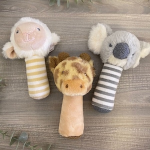 May include: Three plush baby toys with rattle handles. The toys are a white lamb, a brown giraffe, and a gray koala.