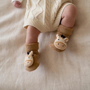 May include: A baby's legs and feet are visible, wearing tan socks with giraffe faces. The baby is also wearing a cream-colored knit romper. The scene is set on a cream-colored fabric.