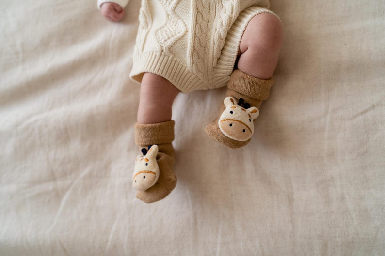 May include: A baby's legs and feet are visible, wearing tan socks with giraffe faces. The baby is also wearing a cream-colored knit romper. The scene is set on a cream-colored fabric.