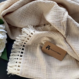 May include: A beige muslin baby blanket with a white crocheted trim and fringe. The blanket has a brown tag with a bear logo and the text "Neutrum Bear".