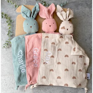 May include: Three personalized bunny security blankets with embroidered names "Charlie", "Grace", and "Oax". The blankets are made of soft, muslin fabric and feature a rainbow pattern.