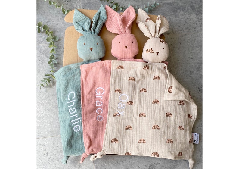 May include: Three personalized bunny security blankets with embroidered names "Charlie", "Grace", and "Oax". The blankets are made of soft, muslin fabric and feature a rainbow pattern.