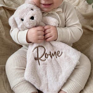 May include: A beige plush security blanket featuring a stuffed animal head with dark eyes and a nose, attached to a soft fabric square. The blanket is embroidered with the name Rome in a brown cursive script, centred on the plush material.