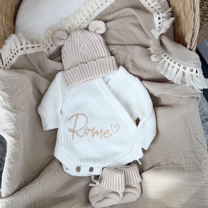 May include: A white long-sleeve knit bodysuit with a wrap-style front featuring the embroidered name Rome and a small heart. The set includes a matching beige knit beanie with bear ears and a pair of beige knit booties with folded cuffs.