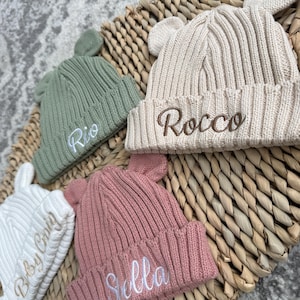 May include: Four ribbed knit beanies with small bear ears on top. The sage green hat features the white embroidered name Rio. The beige hat displays the brown embroidered name Rocco. The pink hat shows the white name Stella. The white hat has text.
