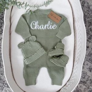 May include: A sage green knitted long-sleeve romper featuring the name Charlie embroidered in white script across the chest. The set includes a matching ribbed beanie with two small bear ears and a pair of soft knitted booties with ribbon ties.