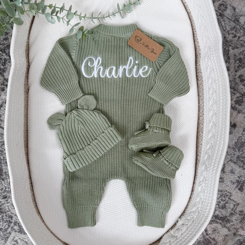 May include: A sage green knitted long-sleeve romper featuring the name Charlie embroidered in white script across the chest. The set includes a matching ribbed beanie with two small bear ears and a pair of soft knitted booties with ribbon ties.