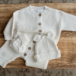 May include: A cream-colored long-sleeve ribbed knit romper featuring a round neckline, a front button placket with light brown buttons, matching ribbed knit mittens, and a cuffed beanie hat with small rounded ears on the top.
