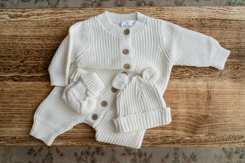May include: A cream-colored long-sleeve ribbed knit romper featuring a round neckline, a front button placket with light brown buttons, matching ribbed knit mittens, and a cuffed beanie hat with small rounded ears on the top.